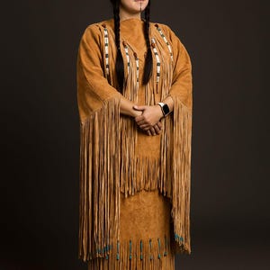 May include: A tan suede dress with long fringe and decorative beading. The dress has a matching fringed shawl. A feather is in the hair. The outfit is worn with tan shoes. The dress is a traditional Native American style.