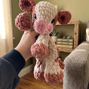 May include: A handmade crochet cow plushie in shades of pink and cream. The plush toy features a pink snout, ears, and hooves, with a cream-colored body and head. The cow is soft and cuddly, perfect for a child's toy or nursery decor.