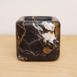 Puede incluir: Un macetero de mármol negro con vetas doradas y blancas. El macetero tiene forma cuadrada con bordes redondeados.