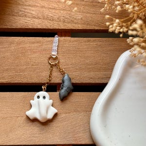 Puede incluir: Un amuleto de fantasma blanco y un amuleto de murciélago negro en una cadena dorada con un soporte de amuleto de teléfono transparente.