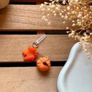 May include: A clear phone dust plug charm with a gold chain, featuring a small orange maple leaf and a miniature pumpkin. The charm is set against a wooden background, evoking an autumn theme.
