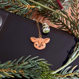 Puede incluir: Un amuleto de ciervo marrón con una cadena dorada y un pequeño amuleto de árbol de Navidad verde. Los amuletos cuelgan de una superficie negra.