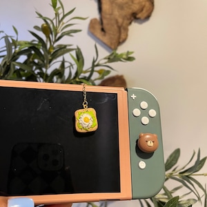 May include: A gold chain with a small avocado toast charm hanging from it. The charm is made of polymer clay and features a green avocado, a white egg, and a brown piece of toast. The charm is hanging in front of a Nintendo Switch console.