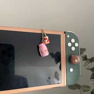 Puede incluir: Un amuleto de té con leche rosa y blanco en miniatura con una cadena dorada y un amuleto de pastel de fresa en miniatura adjunto. Los amuletos cuelgan de una Nintendo Switch.
