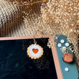 May include: A gold chain with a charm shaped like a cookie with a red heart-shaped jelly filling and white powdered sugar on top. The charm is hanging from a Nintendo Switch console.