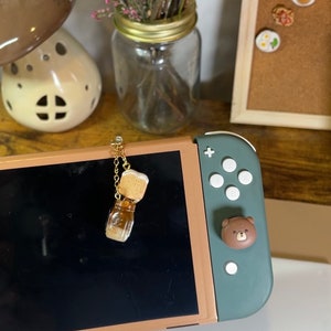 May include: A gold-toned Switch console with a green controller. A small glass bottle charm with a cork stopper hangs from the console. The background includes a vase of dried flowers and a corkboard with decorative pins.