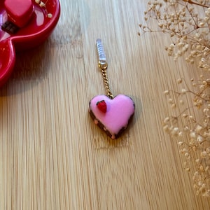 May include: A pink heart-shaped charm with a chocolate border and a small red strawberry on top. The charm is attached to a gold chain and a clear plastic loop.