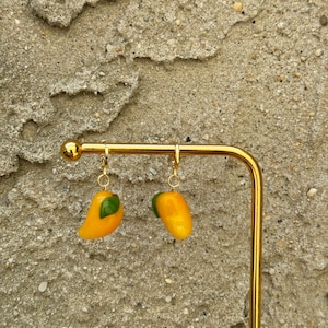 May include: A pair of gold dangle earrings featuring a yellow mango design with a green leaf accent.