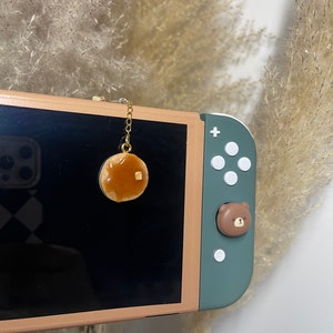 May include: A gold chain with a small pancake charm hangs from a Nintendo Switch console. The console is green and has a brown bear character on the joystick.
