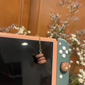 Pode incluir: Um console Nintendo Switch rosa e verde com um amuleto de urso marrom preso ao joy-con direito. Uma corrente dourada com uma pilha de três donuts de argila marrom pendura na parte superior do console.