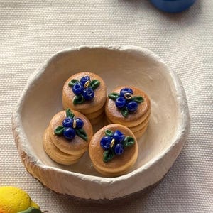 以下が含まれることがあります： 小さなオフホワイトのセラミックボウルには、それぞれ青いブルーベリーと緑の葉で飾られた4つのミニパンケーキが入っています。さらに2つのブルーベリーの彫刻も見えます。パンケーキは積み重ねられており、薄茶色です。