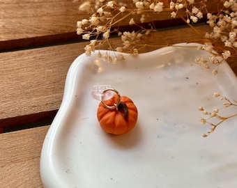 Könnte beinhalten: Ein kleiner orangefarbener Kürbis-Anhänger mit goldenem Stiel, der an einem durchsichtigen Ring an einem weißen Teller hängt.