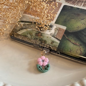 May include: A pink flower charm with green leaves and a gold accent. The charm is attached to a clear phone case.