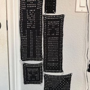 Könnte beinhalten: Eine Sammlung schwarzer, offen gewebter Häkelpaneele in verschiedenen Größen, angeordnet an einer weißen Wand. Die Paneele sind rechteckig und quadratisch, mit einem gitterartigen Muster. Kleine, klare Perlen sind an den Ecken jedes Paneels befestigt.