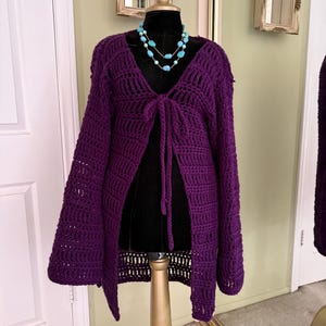 May include: A deep purple, open-front crochet cardigan with long sleeves and a tie closure. The cardigan is displayed on a black mannequin, paired with a turquoise beaded necklace. The garment is handmade and has a textured, open weave design.