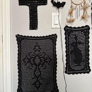 Peut inclure: Décorations murales noires en crochet, comprenant une croix, des chauves-souris et deux pièces rectangulaires aux motifs complexes. Un attrape-rêves avec des plumes et des perles est également visible. Les objets sont exposés sur un mur blanc.