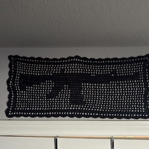 May include: Black crochet wall hanging featuring the silhouette of a rifle. The rectangular piece has a scalloped edge and is mounted on a white wall. The handmade item is a decorative piece.