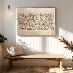 Puede incluir: Una obra de arte enmarcada con caligrafía árabe en escritura marrón oscuro sobre un fondo beige. La obra cuelga sobre un banco de madera con un asiento tejido, complementado con un cojín beige. La escena se desarrolla contra una pared blanca con paneles.