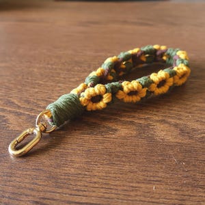 May include: A macrame wristlet keychain featuring a repeating sunflower pattern crafted from yellow, dark green, and brown cord. The loop is secured with a solid green wrapped knot and includes a polished gold-tone metal lobster clasp attachment.