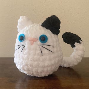 May include: White crocheted cat plush toy with black ears and tail. The cat has blue eyes and a pink nose.