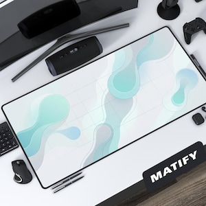 May include: A large rectangular desk mat with a black border and a white surface featuring a teal and gray abstract design. The word "MATIFY" is printed on a black rectangle. Various tech accessories surround the mat.