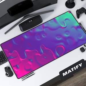 Puede incluir: Una alfombrilla de escritorio rectangular con un diseño abstracto vibrante en tonos de rosa, morado y verde azulado. La alfombrilla tiene una superficie lisa y un borde negro. La palabra "MATIFY" está impresa en una etiqueta negra en la esquina inferior derecha. Un teclado, un ratón y otros accesorios tecnológicos rodean la alfombrilla.