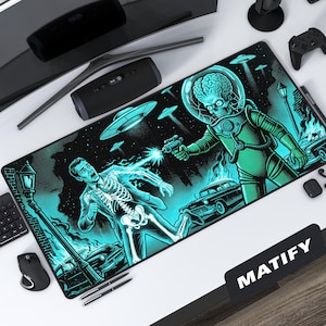 Puede incluir: Alfombrilla de ratón rectangular negra y verde azulado con una ilustración retro de ciencia ficción de un marciano atacando a un hombre con una pistola de rayos. El diseño incluye platillos volantes, coches y un fondo de ciudad. La palabra "MATIFY" está impresa en la parte inferior derecha.