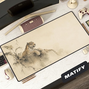 Op de afbeelding: Een rechthoekige beige muismat met een zwarte rand. Het heeft een aquarel illustratie van een luipaard die op een rots rust. Het woord "MATIFY" is in het wit gedrukt op een zwarte rechthoek rechtsonder.