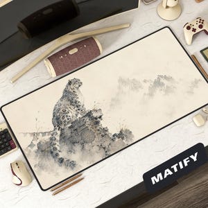 May include: A rectangular desk mat featuring a watercolor illustration of a snow leopard perched on a rocky outcrop, with a misty mountain backdrop. The mat has a black border and the word "MATIFY" is printed on the bottom right.