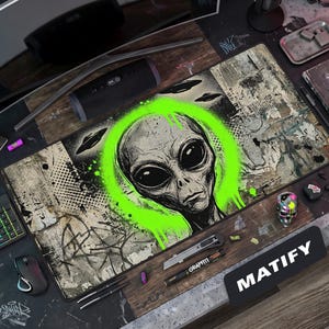 May include: A rectangular desk mat featuring a black and white sketch of an alien head centered within a vibrant neon green circular paint splatter design. The background includes distressed textures, graffiti-style markings, and silhouettes of flying saucers.