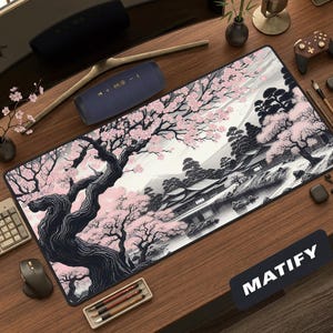 A premium oversized desk mat on a warm wooden desk, featuring a traditional Japanese landscape in an ink-wash and woodblock style. An ancient, gnarled cherry tree with blooming pink sakura blossoms frames the left, contrasting against a monochromatic black, white, and grey landscape with a traditional house near water, pine trees, and a distant Mount Fuji. The setup is styled with a curved monitor, cream mechanical keyboard, wireless controller, and minimalist neutral desk accessories.