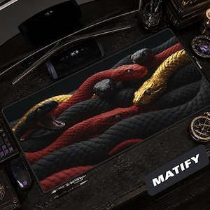 May include: A rectangular desk mat featuring a detailed graphic of intertwined snakes in black, red, and gold. One black snake has its mouth open, while a prominent gold snake is positioned near the top right. The word MATIFY is printed in white.