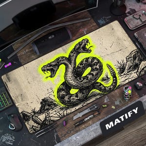 May include: A rectangular desk mat featuring a black and white ink-style illustration of a two-headed rattlesnake with a vibrant neon yellow outline. The background has a distressed, textured beige appearance with desert rock formations.
