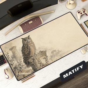 Op de afbeelding: Een rechthoekige beige muismat met een zwarte rand heeft een gedetailleerde illustratie van een bruine uil die op een stenen zuil zit. De achtergrond toont oude ruïnes. Het woord "MATIFY" is gedrukt op een zwarte rechthoek.