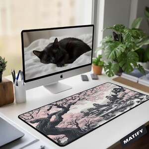 Op de afbeelding: Een rechthoekige bureaumat met een zwart-witte inkttekening van een landschap met roze kersenbloesembomen en een gebouw. De mat ligt op een wit bureau voor een computermonitor waarop een slapende zwarte kat te zien is.