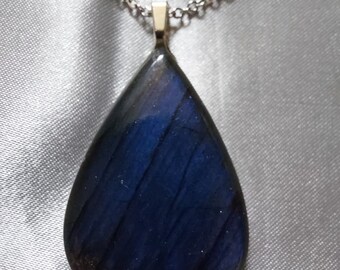 May include: A teardrop-shaped pendant necklace with a silver chain. The pendant is a deep blue color with streaks of lighter blue and a hint of gold. The pendant is made of a polished stone or resin.
