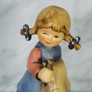 Könnte beinhalten: Vintage Figur einer jungen Frau mit blonden Zöpfen, die ein blaues Kleid, eine gelbe Schürze und braune Schuhe trägt. Sie hält einen Holzstab und hat einen fröhlichen Ausdruck. Die Figur steht auf einem runden, cremefarbenen Sockel.