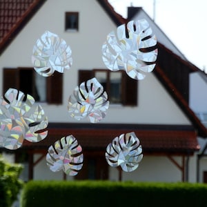 Könnte beinhalten: Ein Fenster mit schimmernden Monstera-Blatt-Aufklebern. Das Fenster befindet sich in einem Haus mit einem roten Ziegeldach.