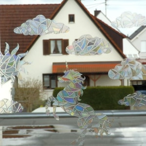 Könnte beinhalten: Ein Stained-Glass-Einhorn-Aufkleber mit schillernden Farben, umgeben von Stained-Glass-Wolken und einem Sonnenaufkleber. Die Aufkleber befinden sich auf einem Fenster mit Blick auf ein Haus.