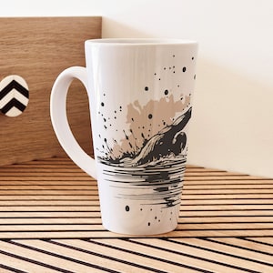 May include: White ceramic mug with a black and brown illustration of a person swimming in a wave. The mug has a handle and is sitting on a wooden surface.