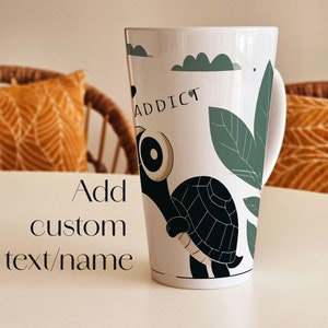 Könnte beinhalten: Weiße Keramiktasse mit einem schwarzen und grünen Schildkröten-Design. Die Tasse trägt den Schriftzug "addict". Fügen Sie einen individuellen Text/Namen hinzu.