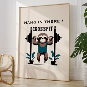 Op de afbeelding: Een ingelijste kunstprint met een cartoon luiaard die gewichten heft. De luiaard is bruin en draagt een turquoise tanktop. De woorden "HANG IN THERE! CROSSFIT" staan boven de luiaard gedrukt. De achtergrond is lichtbeige.