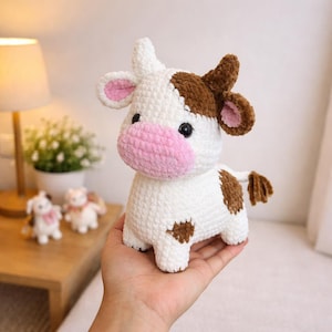 May include: A handmade crochet cow plushie. The cow is white with brown spots and accents, including the ears and tail. It has a pink muzzle and inner ears, and black safety eyes. The plushie is held in a hand.