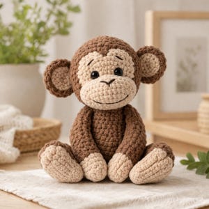 Può includere: Una scimmia all'uncinetto fatta a mano nei toni del marrone e beige. Il peluche ha una faccia sorridente, grandi orecchie ed è seduto su una superficie bianca. La scimmia è un giocattolo morbido e coccoloso.