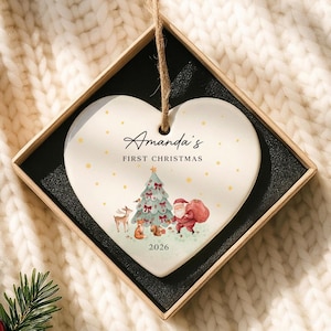 May include: A heart-shaped white ceramic ornament featuring a watercolor illustration of a Christmas tree, Santa Claus, and forest animals. The text reads Amanda's FIRST CHRISTMAS 2026. It hangs by a jute twine string inside a square gift box.