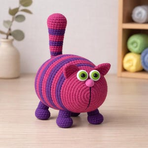 May include: A handmade crochet cat in shades of pink and purple. The cat has large, green-eyed, a pink nose, and a striped tail. The cat is standing on four purple legs. This whimsical cat is a unique decorative item.