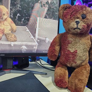 May include: A brown teddy bear with red markings, featuring a stitched black eye and a sorrowful expression. Another teddy bear is visible on a computer monitor in the background.