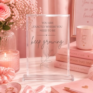 May include: A rectangular clear acrylic plaque featuring the printed text "YOU ARE EXACTLY WHERE YOU NEED TO BE" followed by a small heart icon and the script text "keep growing" above a line drawing of a leafy botanical branch.