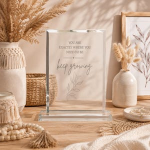 May include: A rectangular glass plaque with beveled edges, featuring the printed text "YOU ARE EXACTLY WHERE YOU NEED TO BE" and "keep growing" above a line drawing of a leafy branch. The plaque is mounted on a clear glass base.
