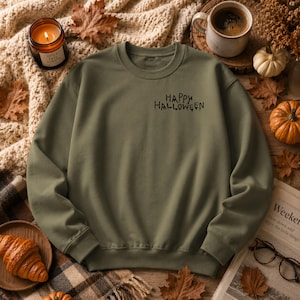 Puede incluir: Una sudadera de cuello redondo en color verde oliva con el texto HAPPY HALLOWEEN impreso en una fuente negra estilizada y goteante en la parte superior izquierda del pecho. La prenda tiene mangas largas, cuello acanalado y puños acanalados.